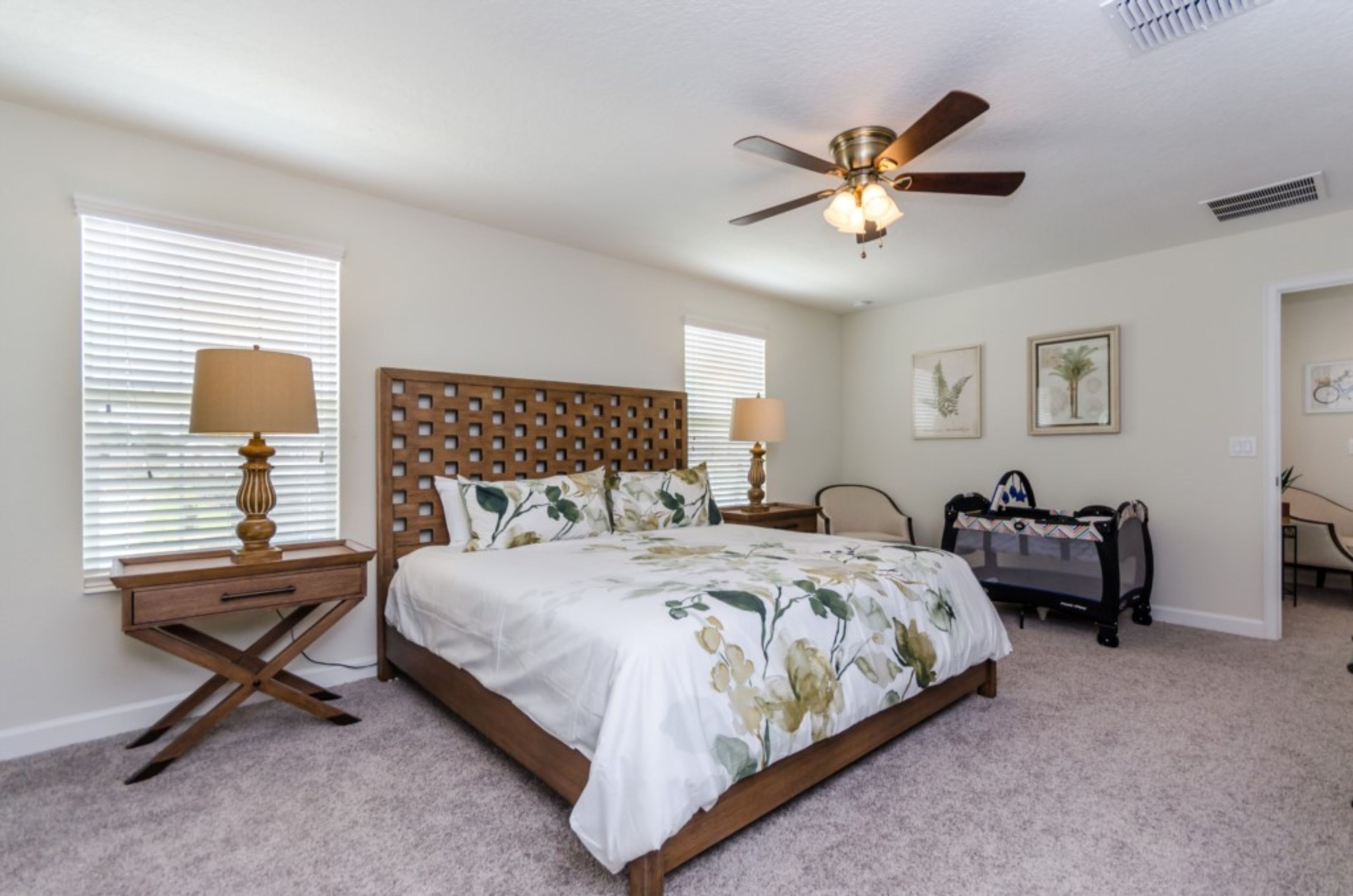 A bedroom with a wooden headboard, a bed with floral bedding, two bedside tables with lamps, a ceiling fan, and a small desk with a chair.