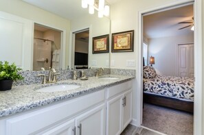 A bathroom with a double sink, a large mirror, and a shower. There is a bed with a patterned bedspread in the adjacent room.