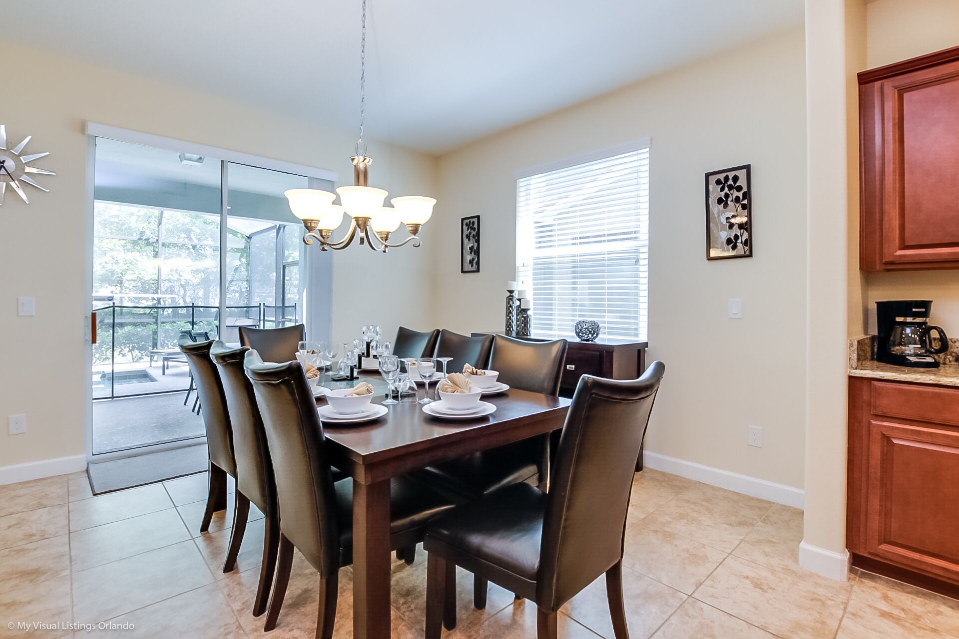A dining area with a large table set for six, surrounded by chairs. There is a chandelier above the table, and a kitchen area is visible to the right.