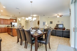 A dining area with a wooden table and chairs, a kitchen in the background, and a living room with a sofa and television.