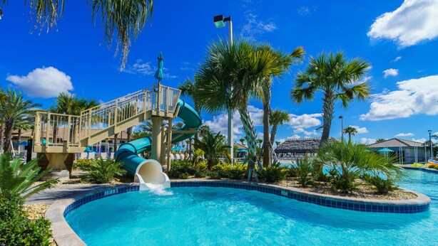 Une piscine avec un toboggan, entourée de palmiers et surplombée d’un ciel bleu azur.