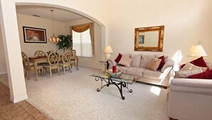 Uma sala de estar com sofá, mesa de centro de vidro, área de jantar com mesa e cadeiras, e um espelho grande na parede.