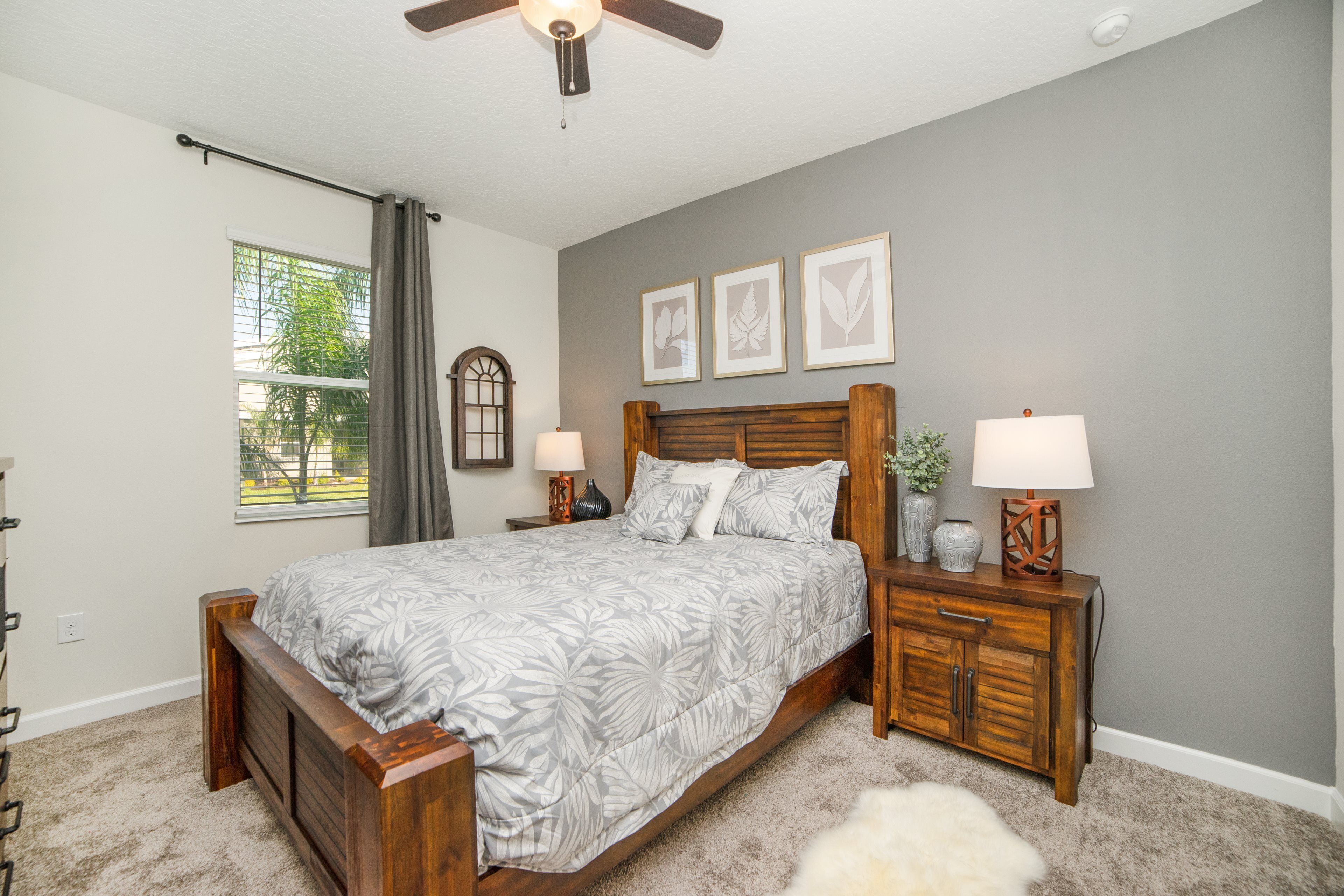 A bedroom with a large bed, two bedside tables with lamps, a ceiling fan, a window with curtains, and three framed pictures on the wall.