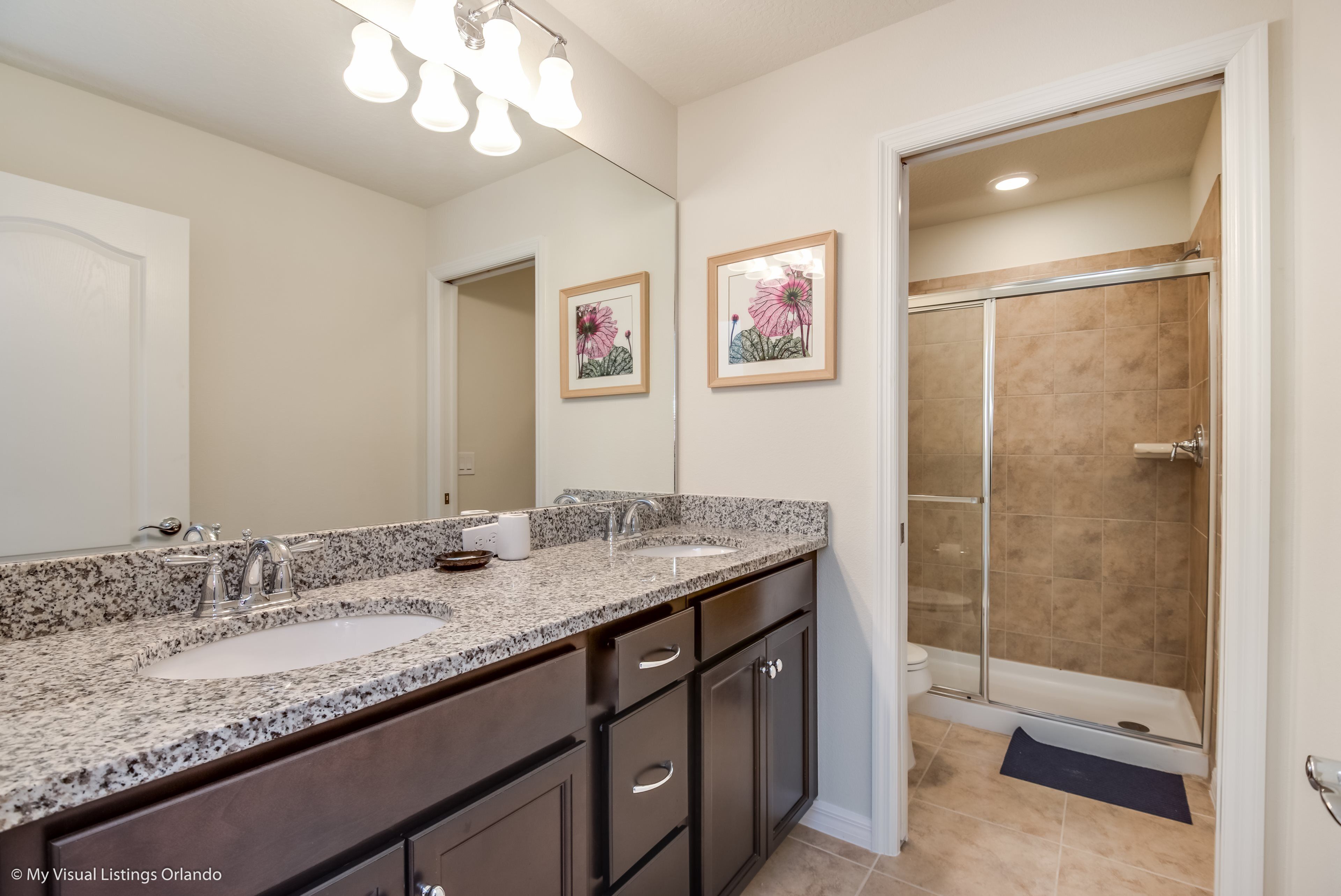 Une salle de bain dotée d’un meuble-lavabo à deux vasques, d’un grand miroir, d’œuvres d’art florales encadrées et d’une douche à l’italienne.