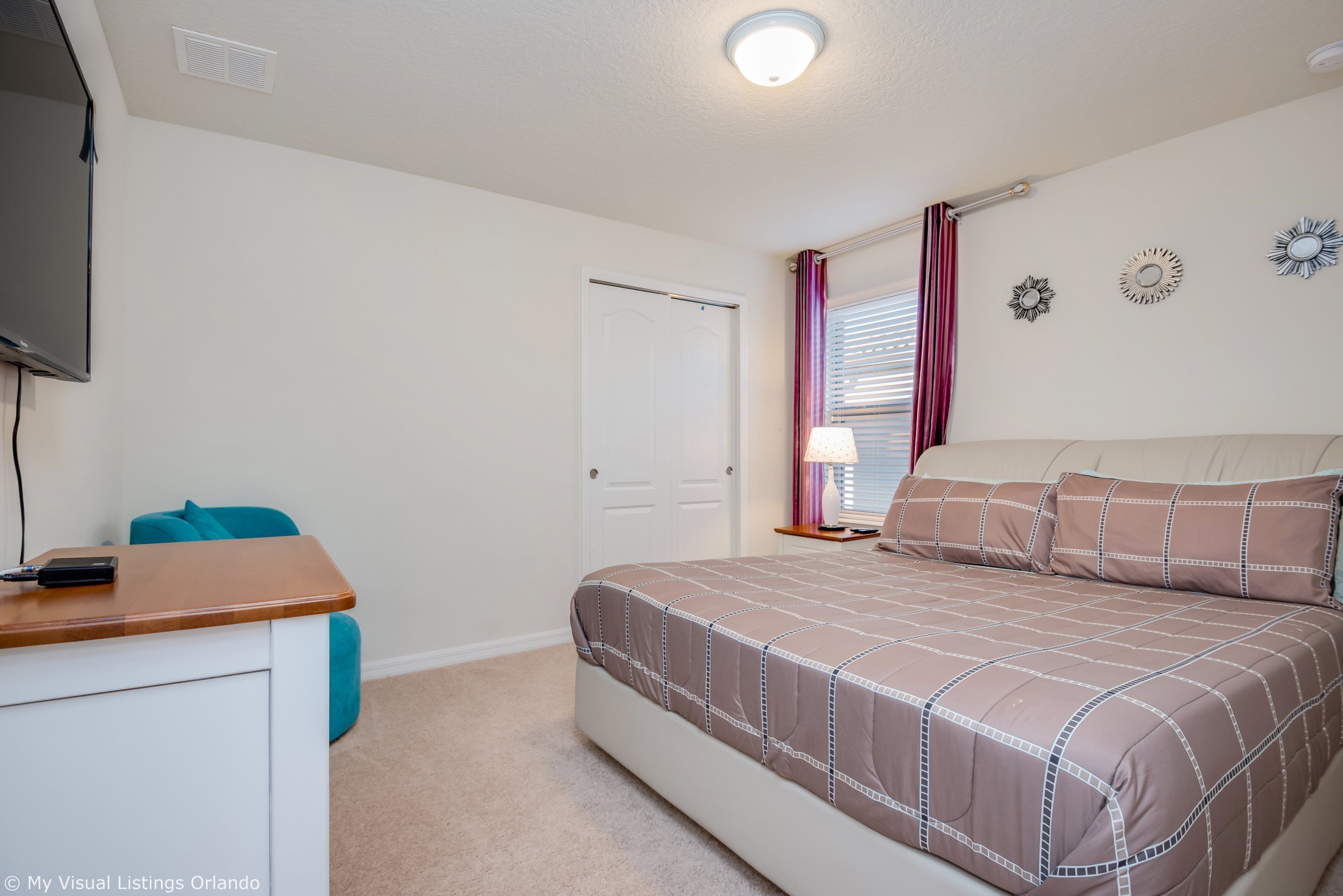 A bedroom with a bed, a desk, a chair, a TV, a door, and a window with curtains.