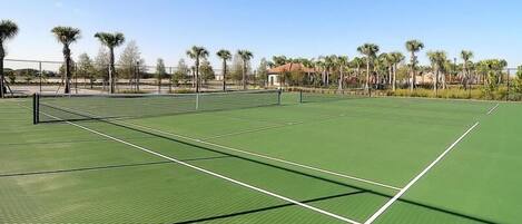 An outdoor tennis court with a green surface, surrounded by palm trees and a clear sky.