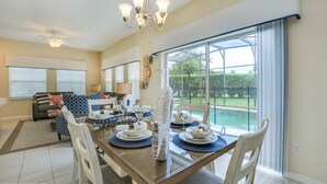 A dining area with a glass table, white chairs, and a chandelier. There is a sofa and a window with blinds in the background. - 4 Bedroom Townhouse with Pool (Kissimmee)