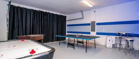 A room with a foosball table, a ping pong table, and a foosball table with a red ball.