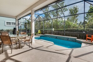 Zona de piscina exterior techada con mesa y sillas, rodeada por un cerco de madera y árboles.