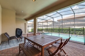 A patio with a wooden table and chairs, a grill, and a view of a swimming pool through a glass enclosure. - 8 Bed Villa with Games, Pool & Spa (Kissimmee)