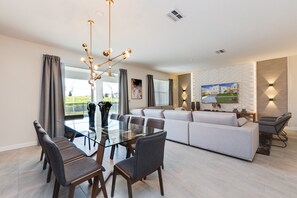 A modern living room with a glass dining table, grey sofas, and a large TV mounted on the wall.