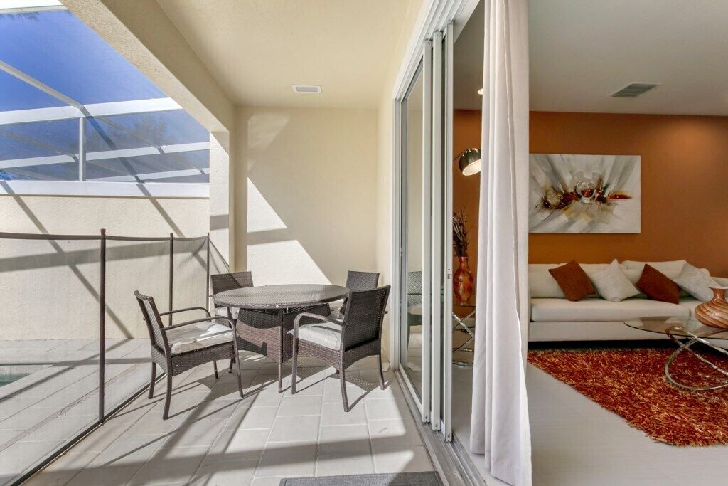 A balcony with a glass door, a round table with chairs, and a sofa with cushions.