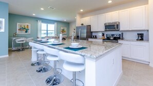 A modern kitchen with white cabinets, a granite countertop, and stainless steel appliances. There are four white bar stools at the island. - Cool and Modern 9 Bed 7 Ba Villa Pool & Spa 6160boa (Davenport)