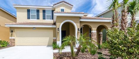 A two-story house with a beige facade, a garage, and a landscaped front yard with palm trees and shrubs.