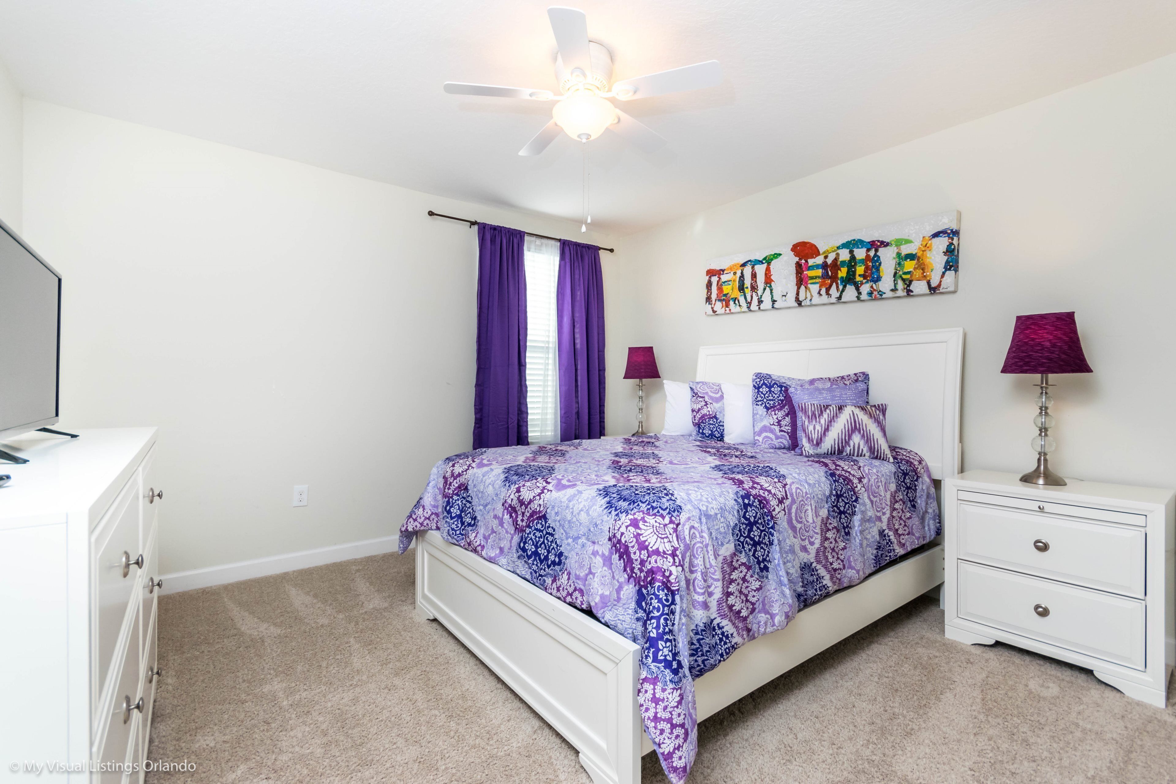 A bedroom with a bed, nightstands, a ceiling fan, a window with purple curtains, and a wall art piece.
