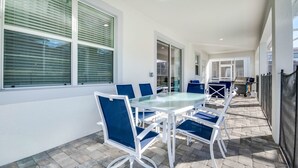 A covered patio with a glass-top table and blue chairs, large windows with blinds, and a paved walkway. - 9085hs (Davenport)