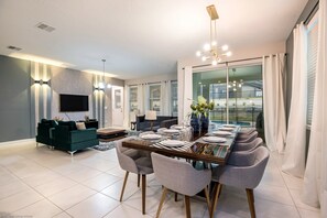 A modern dining area with a long table set for six, surrounded by grey chairs. A dark green sofa and a small side table with a lamp are visible in the background. - 1780spp New 7 Bedroom 5 Bathroom Solara Villa (Kissimmee)