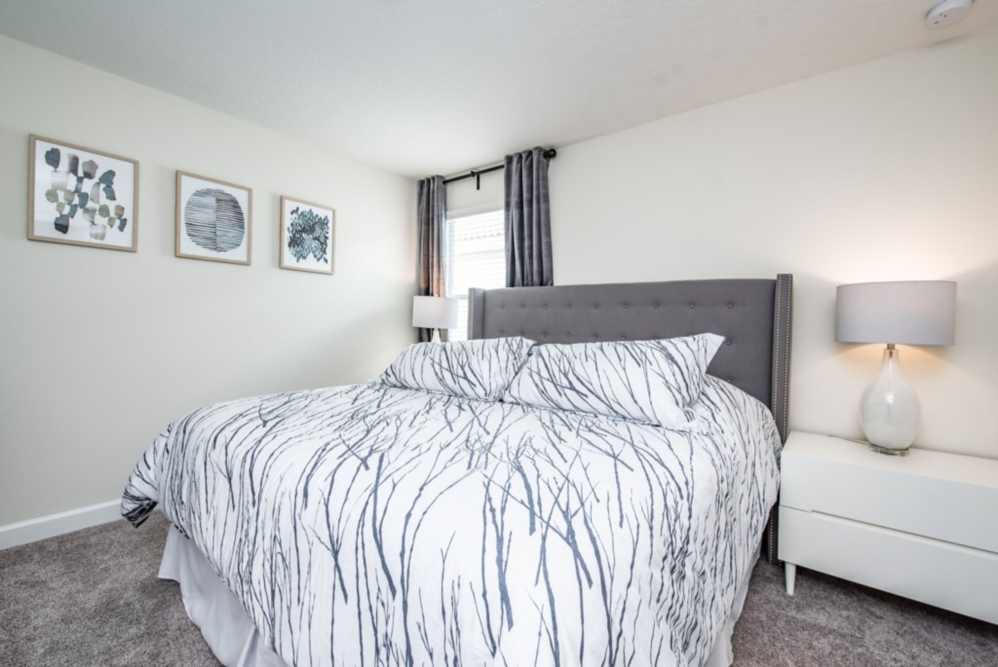 A bedroom with a large bed, a bedside table with a lamp, and three framed artworks on the wall.