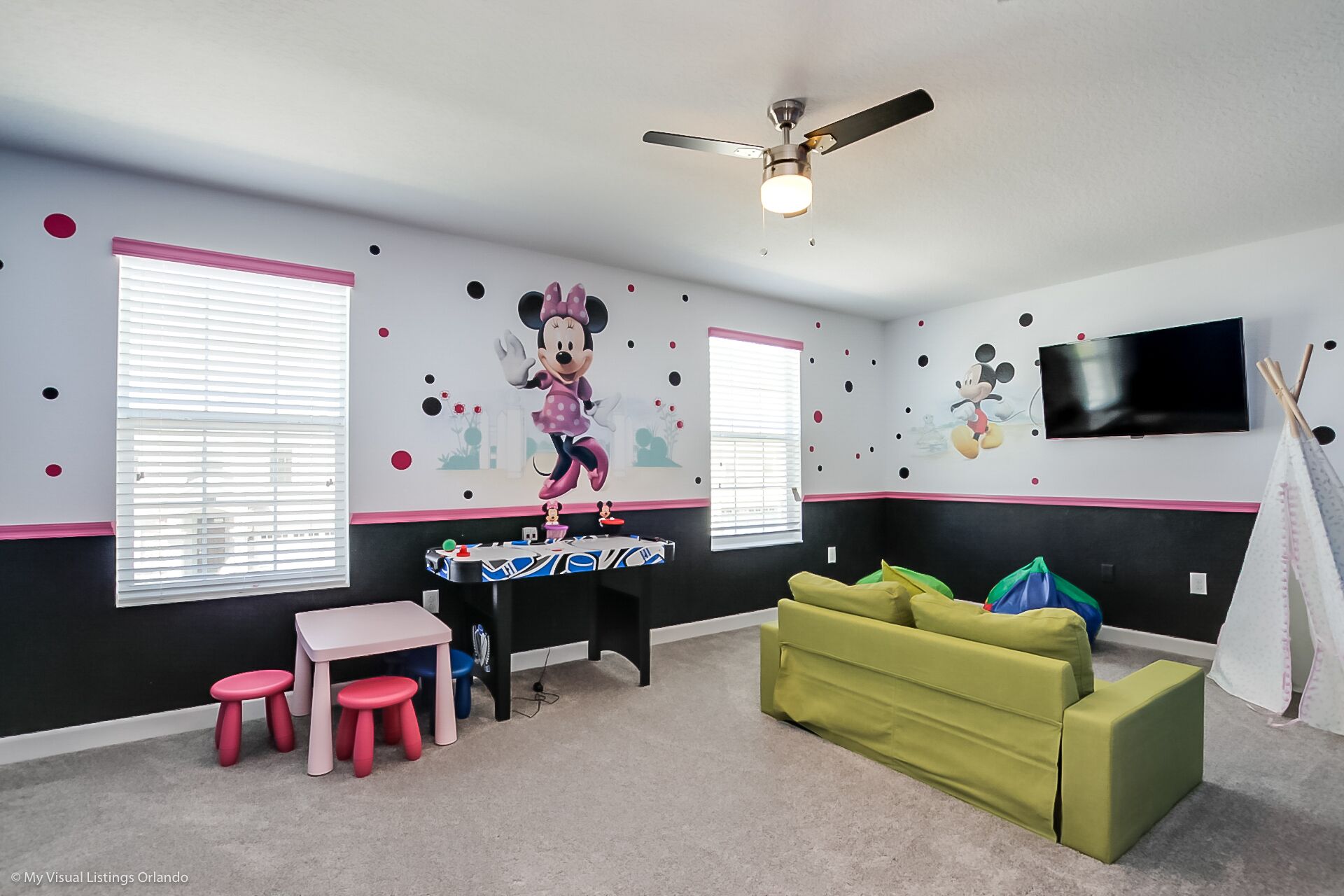 Une salle de jeux pour enfants avec un autocollant mural à l’effigie de Minnie Mouse, un ventilateur de plafond, un téléviseur et un canapé vert.