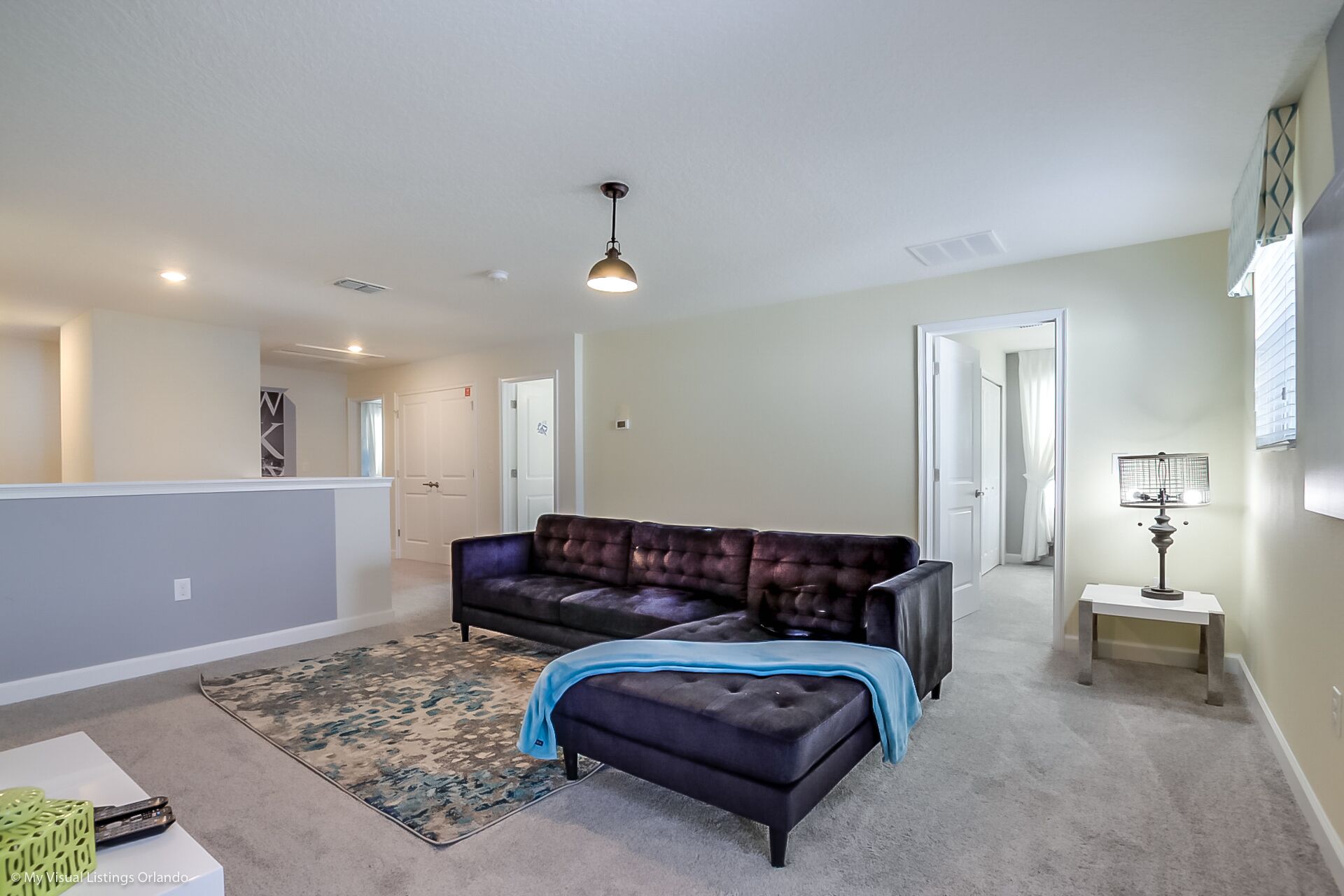 A spacious living area with a dark brown leather sofa, a matching ottoman, and a blue throw blanket. There is a small side table with a lamp, a patterned rug, and a view of a hallway leading to another room.