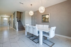 A modern dining area with a white table and chairs, a chandelier, and a staircase in the background. - 8911sd Champions Gate 8 Bedroom Resort Villa (Davenport)