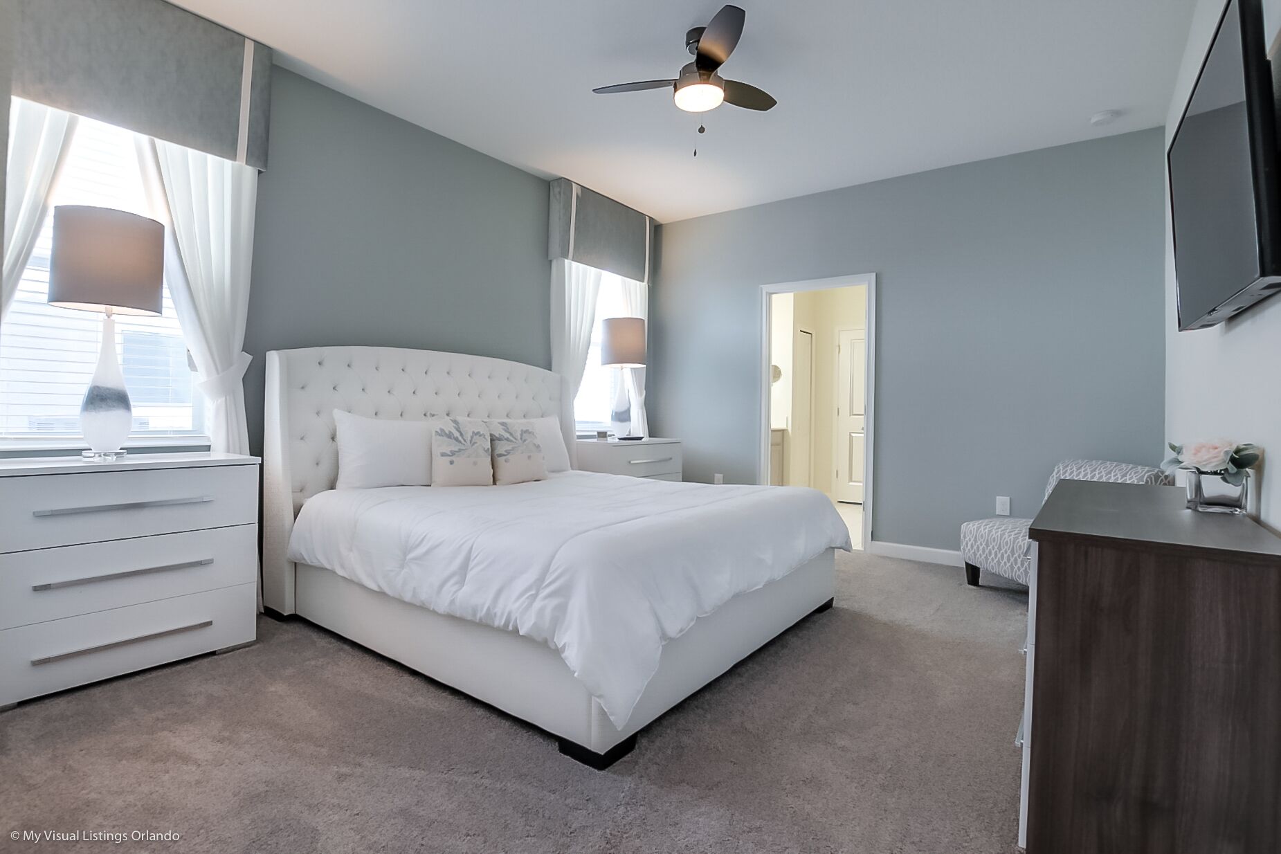 A bedroom with a large bed, a ceiling fan, a dresser, and a TV mounted on the wall.