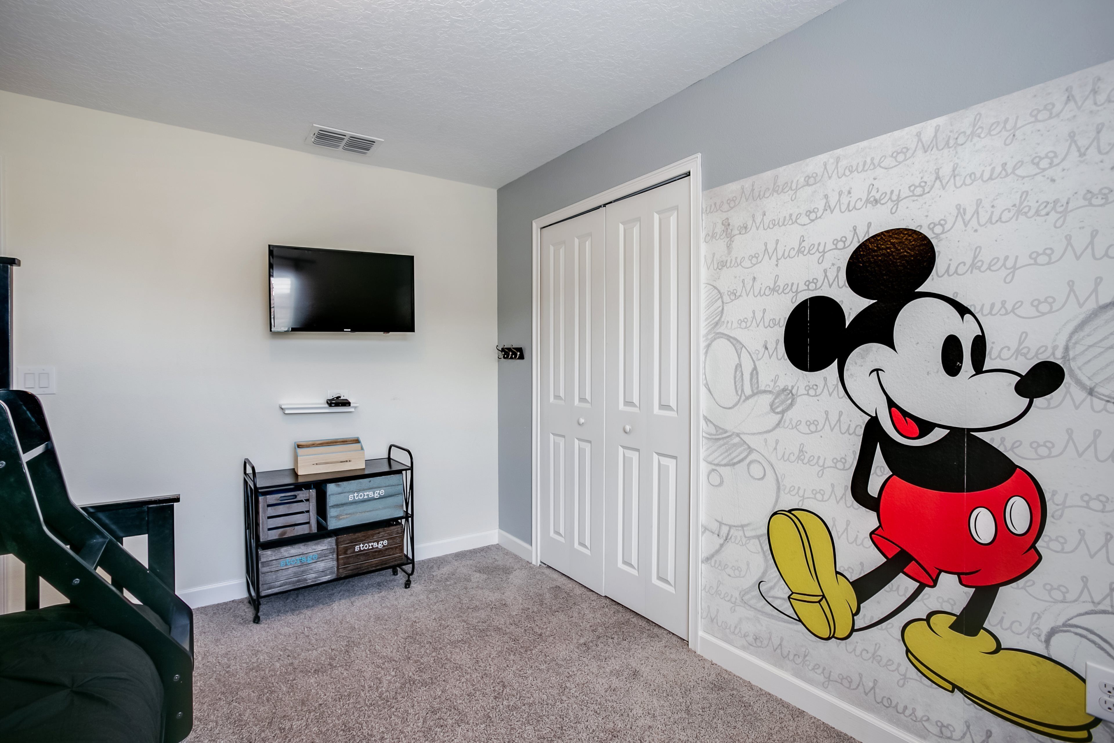 A room with a wall mural of Mickey Mouse, a flat-screen TV mounted on the wall, a storage unit with labeled boxes, and a closed door.