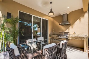 Een terras buiten met een glazen tafel, rieten stoelen, een barbecue en potplanten.