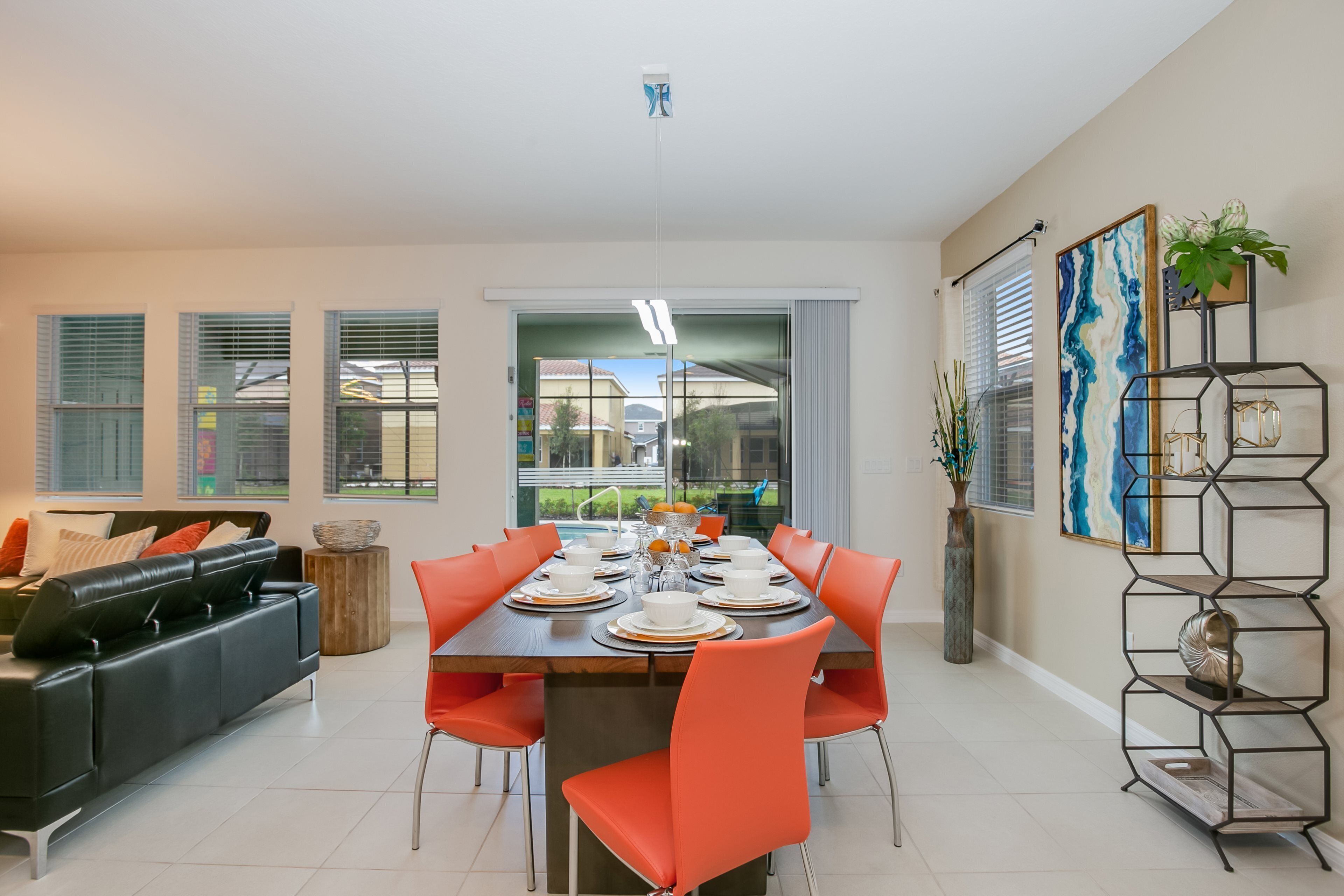 Un comedor moderno con una gran mesa dispuesta para seis personas, rodeada de sillas anaranjadas. Hay un sofá de cuero negro y una mesita de madera con un tazón. Una estantería metálica con objetos decorativos está a la derecha.