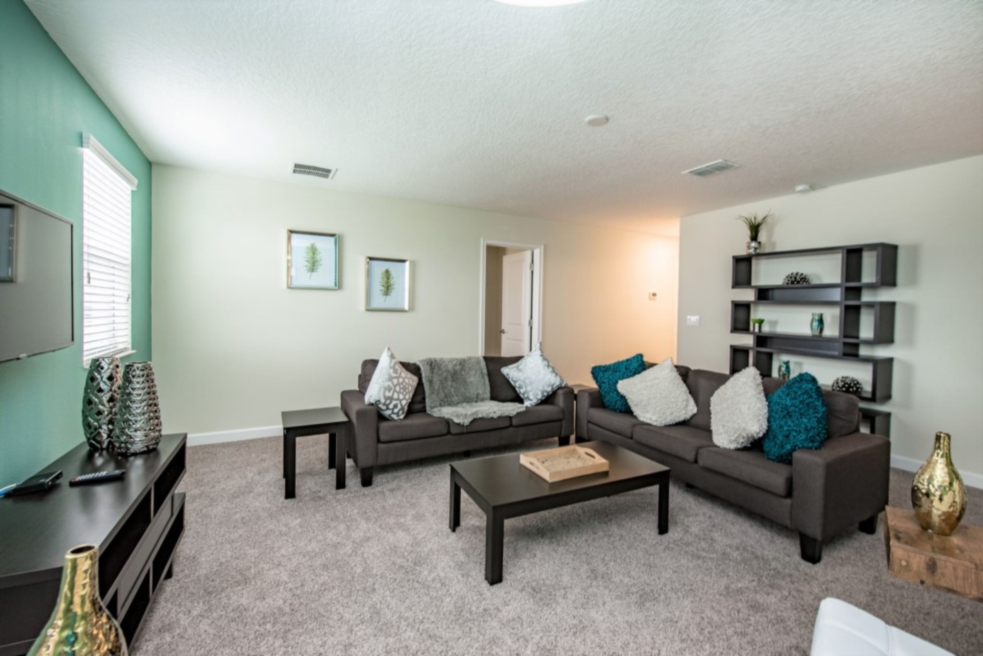 A living room with a sectional sofa, a coffee table, and a TV on a stand. There are two framed pictures on the wall and a bookshelf with decorative items.