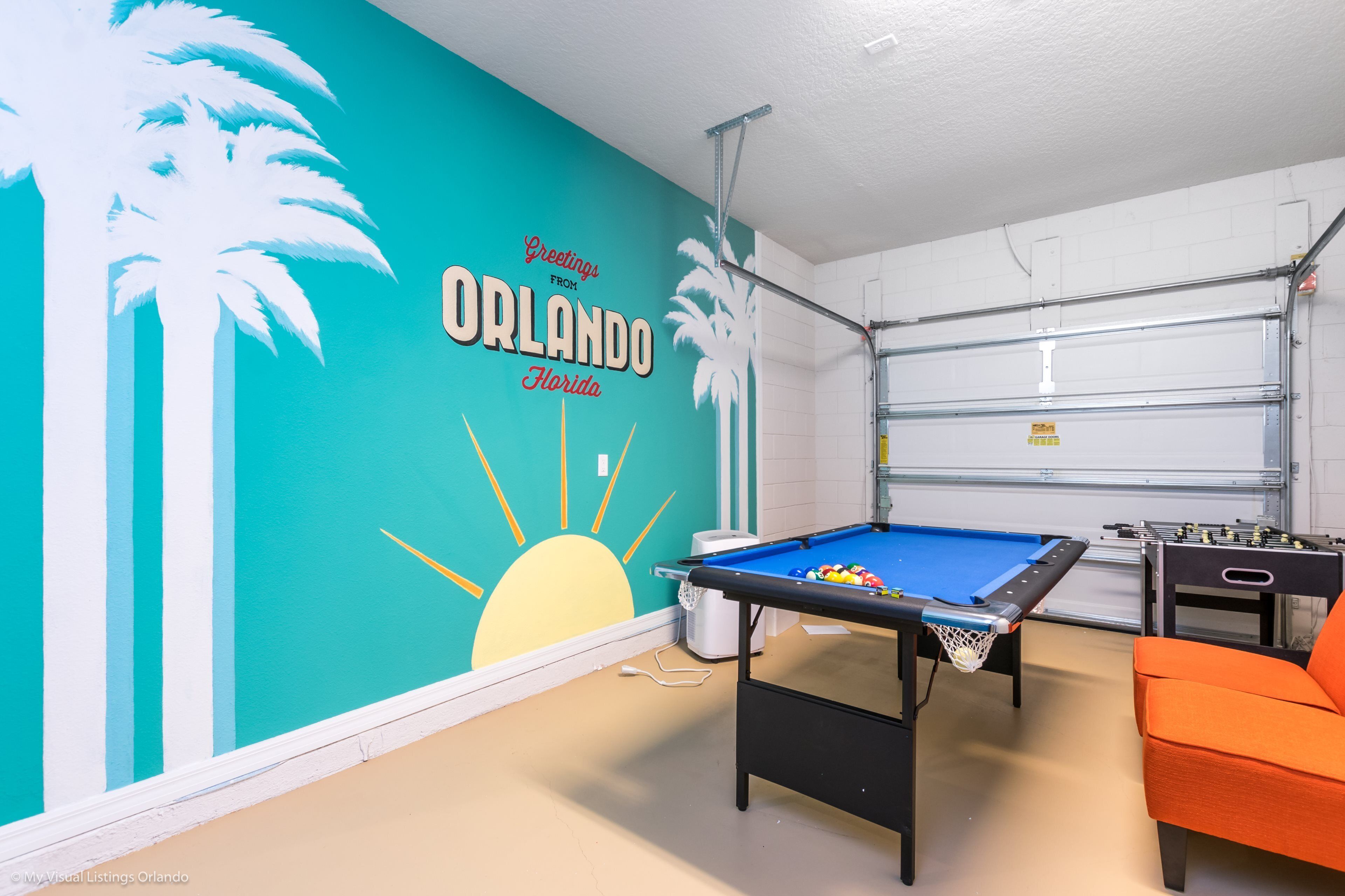 A room with a pool table, a foosball table, and a mural of palm trees and a sun.