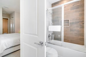 A hotel bathroom with a shower, toilet, and a bed in the adjacent room.