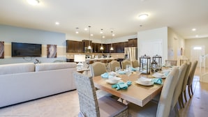 A modern kitchen with dark cabinets, a dining area with a wooden table set for six, and a living area with a sofa and TV. - 9 Bed Villa with Games, Pool & Spa (Kissimmee)