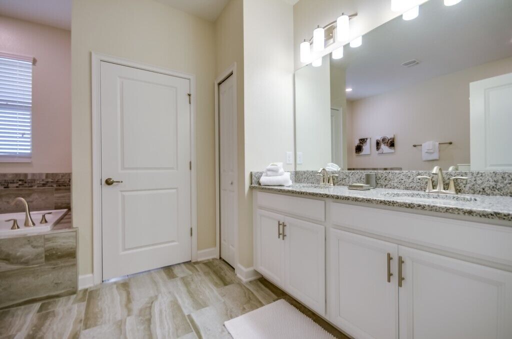 A bathroom with a white door, a sink with a faucet, a mirror with lights, and a countertop with a towel.