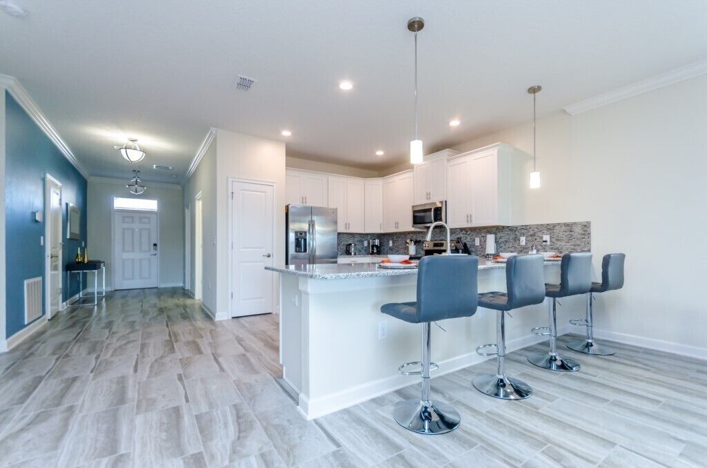 A modern kitchen with white cabinets, a granite countertop, and stainless steel appliances. There are four bar stools and a pendant light above the island.