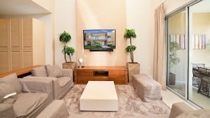 A modern living room with a flat-screen TV mounted on the wall, a wooden entertainment unit, beige sofas, a white coffee table, and two potted plants. - New Magic Villa Resort Townhome 3123bra (Kissimmee)