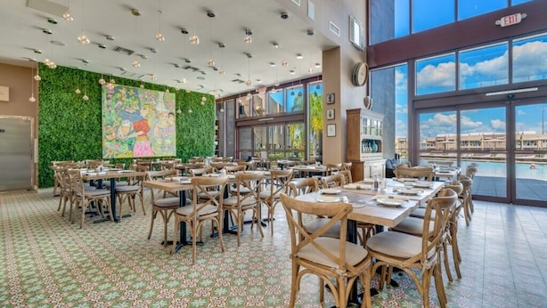A spacious dining area with tables and chairs, a green wall with artwork, and large windows offering a view of buildings and a body of water. - New Magic Villa Resort Townhome 3123bra (Kissimmee)