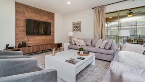 A modern living room with a flat-screen TV mounted on a wooden wall, a grey sofa, a white coffee table, and a large window with curtains. - 8 Bed 6 Ba Villa with Games, Pool & Spa (Kissimmee)