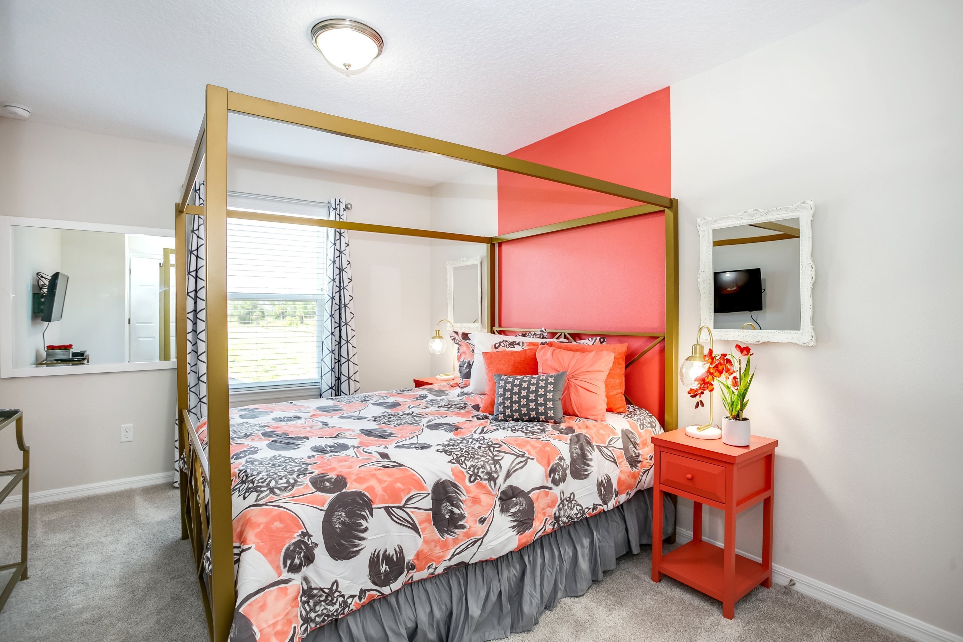 A bedroom with a four-poster bed, a red accent wall, a bedside table with a lamp, and a mirror.