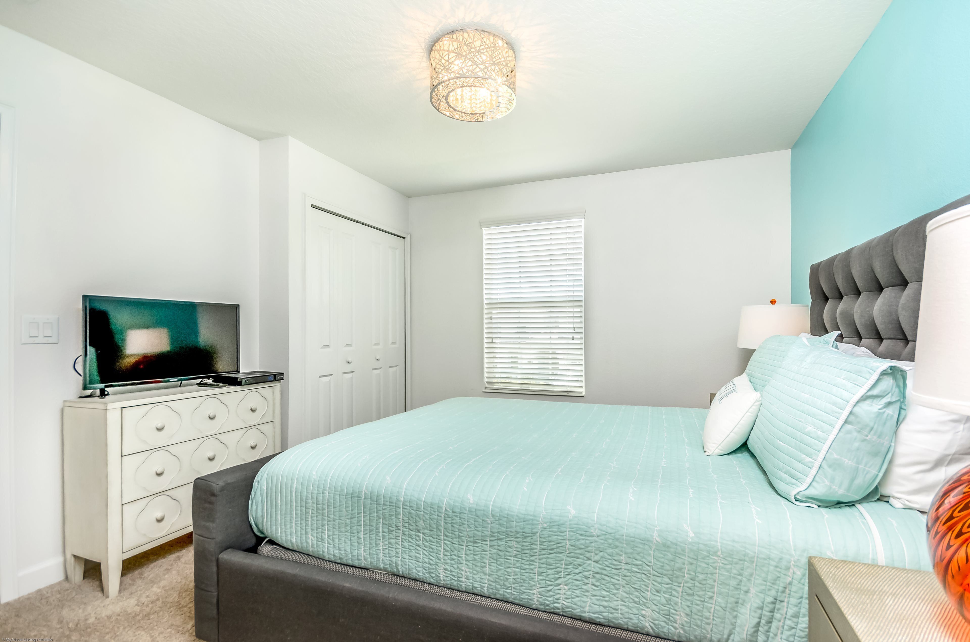 Een slaapkamer met een bed, een dressoir, een televisie, een raam met zonwering en een plafondlamp.