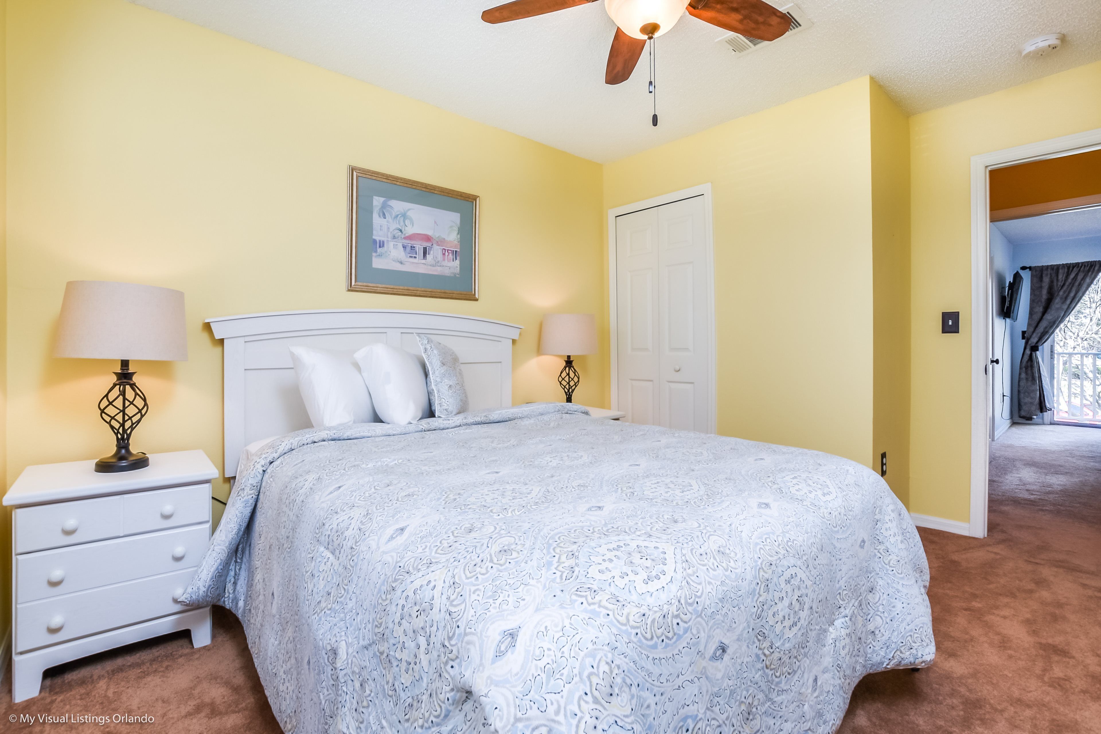 A bedroom with a white bed, two bedside tables with lamps, a ceiling fan, a door, and a framed picture on the wall.