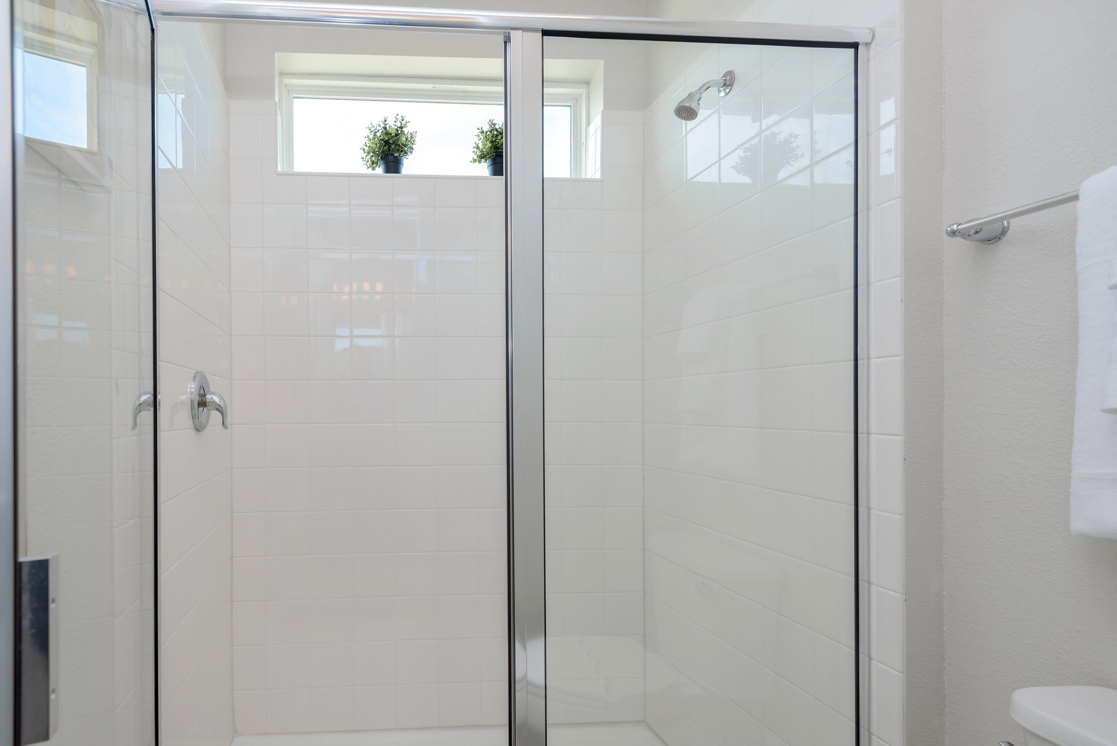 A modern bathroom with a glass shower enclosure, white tiles, and a visible window with plants.