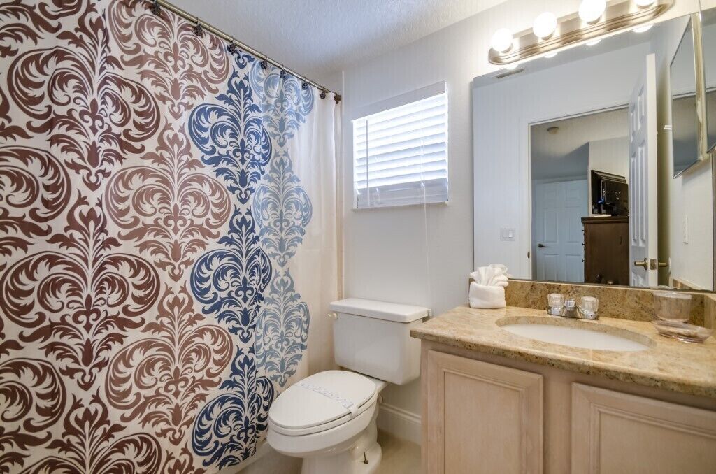A bathroom with a patterned shower curtain, a toilet, a sink with a mirror, and a window with blinds.