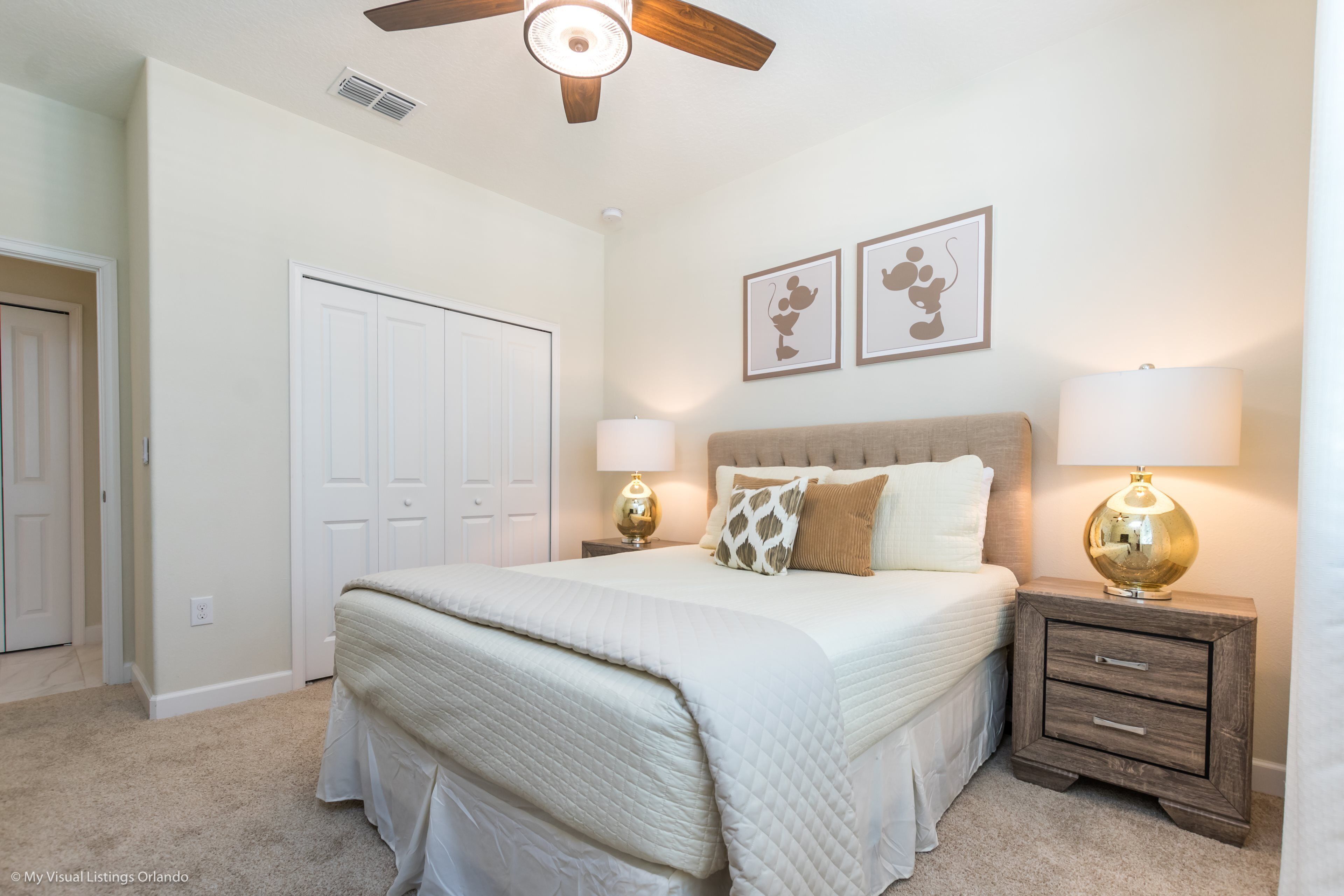 A bedroom with a large bed, two bedside tables with lamps, a ceiling fan, and two framed pictures on the wall.