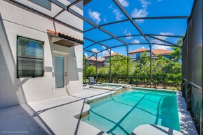 A swimming pool with a glass enclosure, a white building with a door and windows, and a view of neighboring houses and trees.