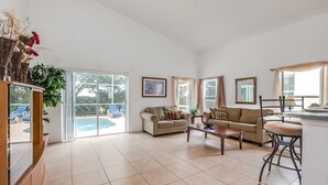 A spacious living room with a beige sofa, a coffee table, and a bar area. There are large windows with curtains, a pool visible outside, and a framed picture on the wall. - 435castle (Davenport)