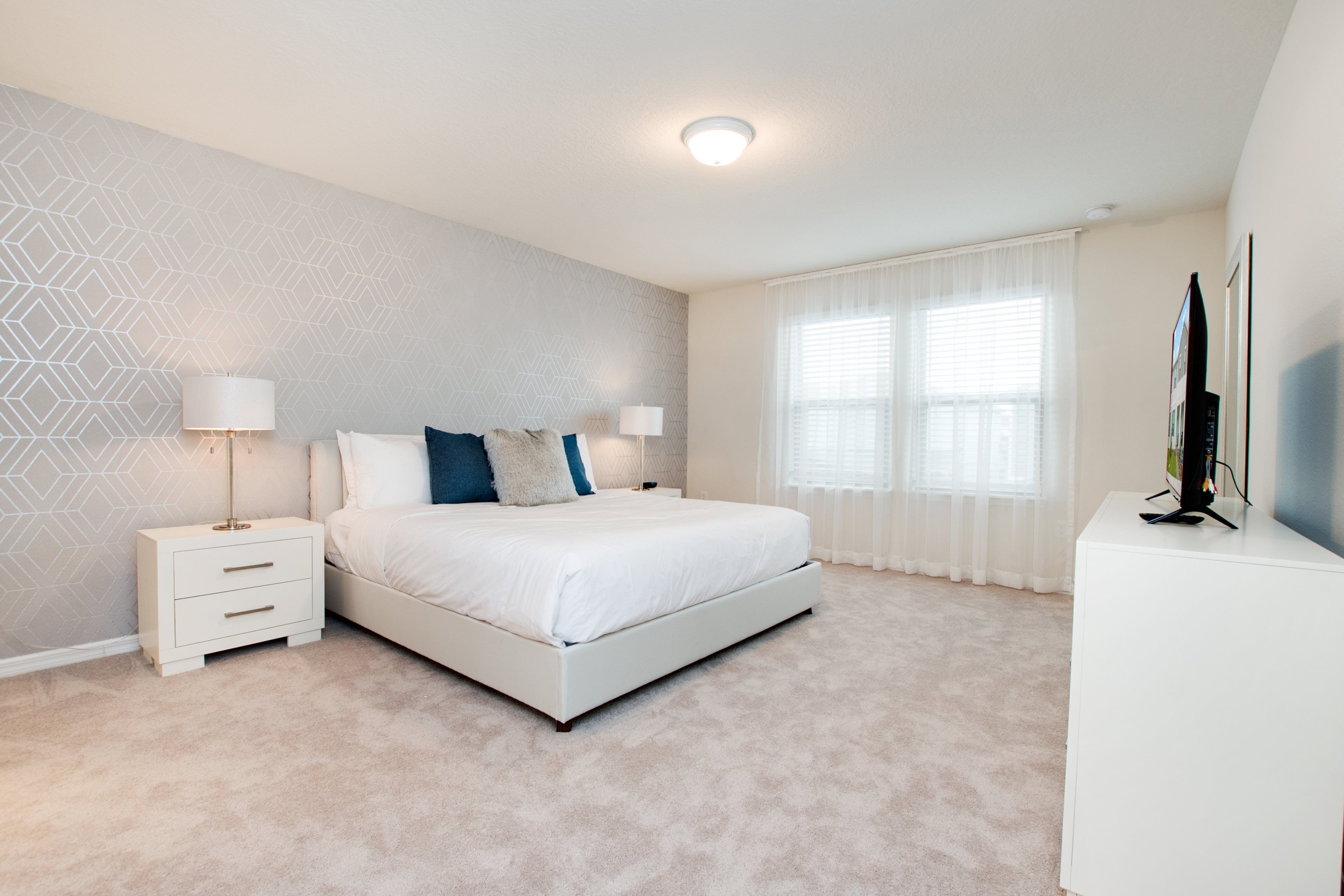 A modern bedroom with a large bed, two bedside tables, a TV, and a window with sheer curtains.
