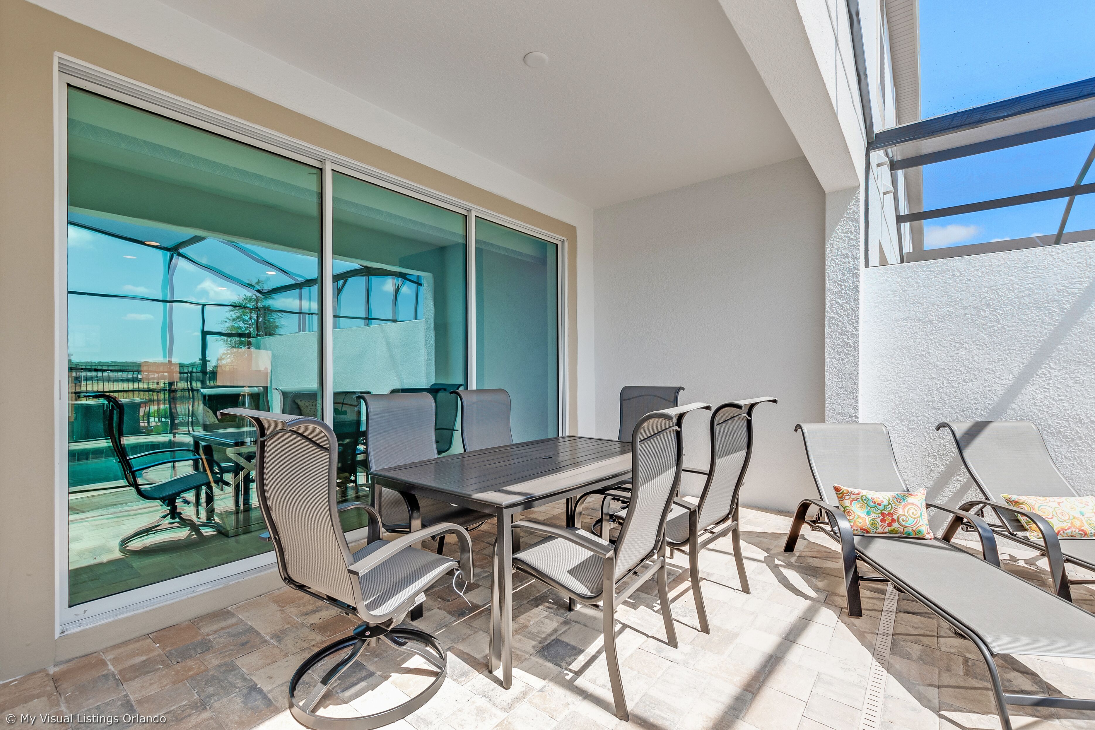 A sunlit patio with a glass wall, a dining table, chairs, and a lounge area.