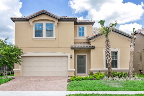 A two-story house with a beige exterior, a white garage door, and a small front yard with a palm tree and shrubs. - 8917bengal Sleeps 20 (Kissimmee)