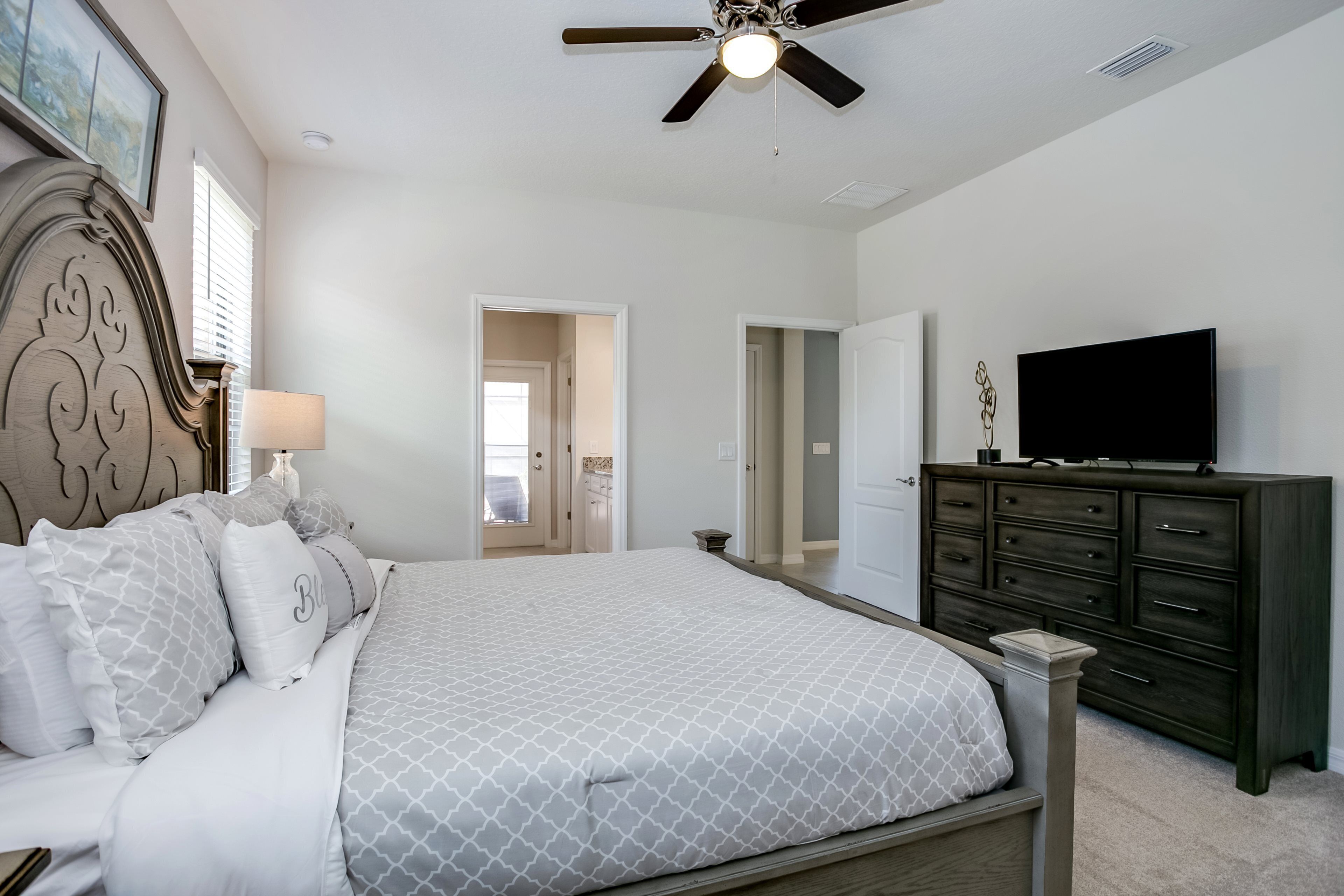 A bedroom with a bed, dresser, TV, ceiling fan, and a door leading to another room.